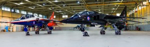 Two SEPECAT Jaguar aircraft, one in red white and blue and one in black, parked nose-to-nose in a hangar