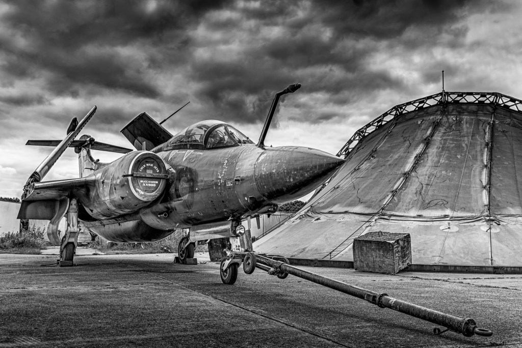 Black and white side view of Blackburn Buccaneer XX900 with a tow bar attached beside a hangar
