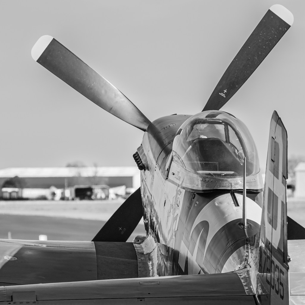 Black and white close-up of the nose and propeller of a P-51D Mustang with a hangar in the background