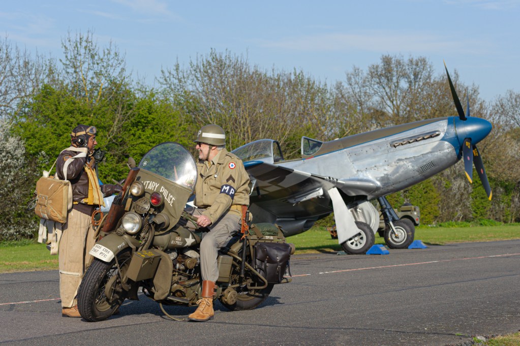 Reenactors in US uniform with a wartime motorcycle in front of a P-51D Mustang at North Weald