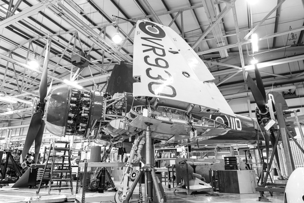Black and white wide-angle view of Hawker Sea Fury VR930 in a maintenance hangar with a wing folded and the serial visible