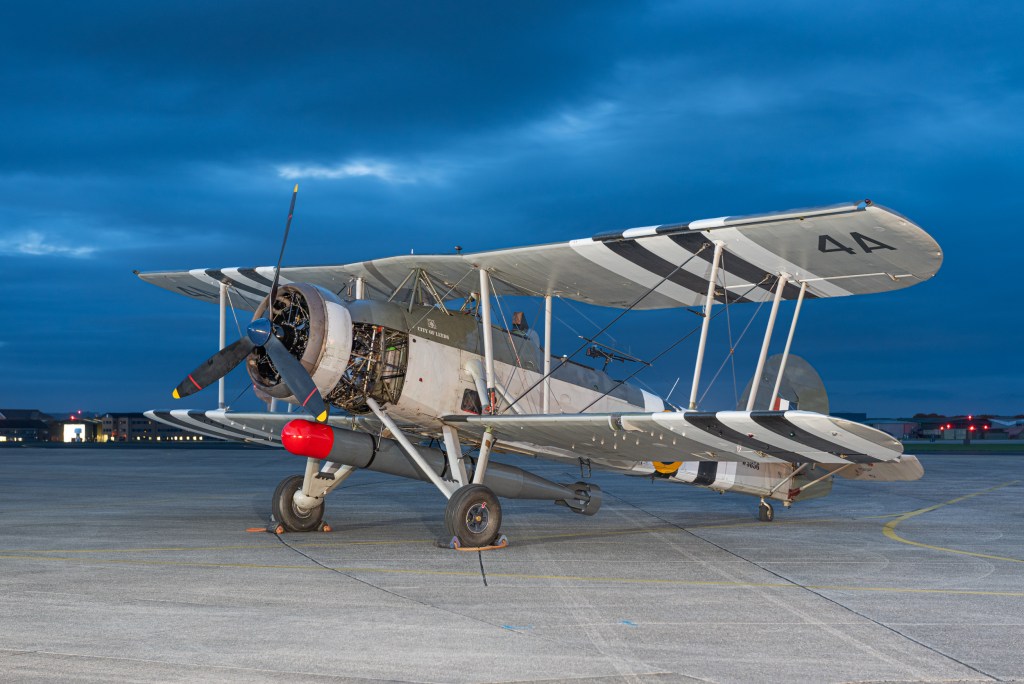 Fairey Swordfish W5856 biplane with black and white invasion stripes on the apron at RNAS Yeovilton under a deep blue twilight sky.
